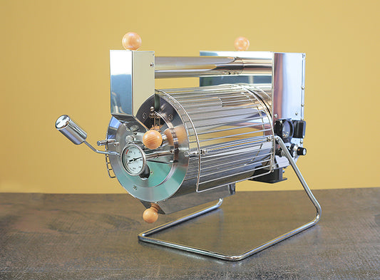 Angled frontview of the Quest M6 electric coffee roaster. The barrel faces forward with a chaff collector below and an air intake tube above. 
